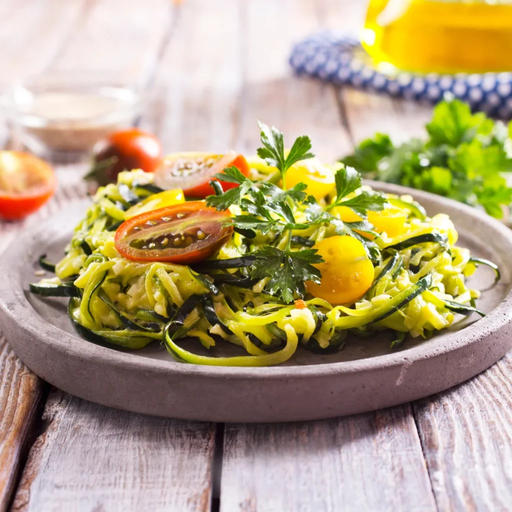 Prato fundo contendo tiras espiralizadas de abobrinha crua envolvidas em um molho pesto verde brilhante com metades de tomate cereja por cima