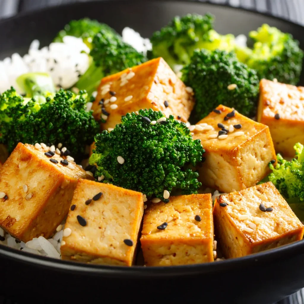 Tigela de cerâmica escura contendo cubos de tofu grelhados com gergelim, acompanhados de arroz e fatias de vagem